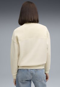 Person iført en cremet langærmet struktureret sweater og lyseblå jeans, stående med ryggen imod kameraet mod en grå baggrund.