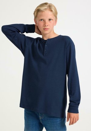 Marineblauwe henley-shirt met lange mouwen, drie knopen bij de kraag, geribbelde textuur en een relaxte pasvorm, gecombineerd met blauwe jeans.