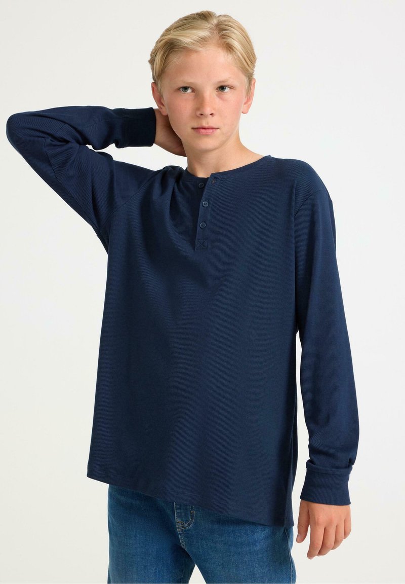 Marineblauwe henley-shirt met lange mouwen, drie knopen bij de kraag, geribbelde textuur en een relaxte pasvorm, gecombineerd met blauwe jeans.