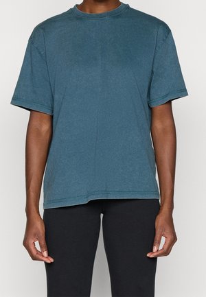 Person iført en kortærmet, falmet blå T-shirt med rund hals og sorte bukser, vist fra nakken til hofterne mod en ensfarvet baggrund.