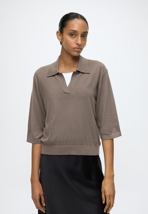 Femme aux cheveux foncés en chignon bas, portant un haut taupe à manches courtes avec col sur une chemise blanche et une jupe noire sur un fond uni.