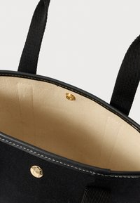Černá taška přes rameno s béžovým interiérem, černými tkanými držadly, zlatým patentním uzávěrem a viditelným bílým prošíváním podél horního okraje.