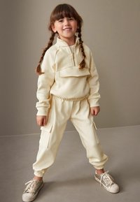 Cremefarvet fleece træningsdragt med lynlås ved halsen, cargo-lommer, elastiske manchetter og parret med glitrende sølv sneakers.