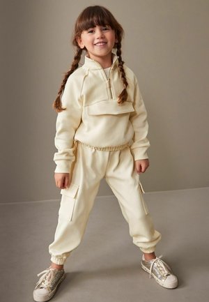 Kreminės spalvos fliso treniruotės kostiumas su užtrauktuku ant kaklo, cargo kišenėmis, elastinėmis rankogaliais ir derinamas su blizgančiais sidabriniais sportbačiais.