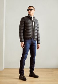 Veste noire rembourrée avec des coutures horizontales, en matériau léger, associée à un jean bleu foncé et des mocassins noirs. Design ajusté.