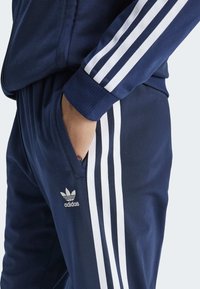 Navy tracksuit featuring white three-stripe design on sleeves and sides. Material appears smooth with a ribbed cuff at the ankle. Adidas logo displayed.