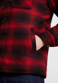 Chemise en flanelle à carreaux rouges et noirs avec patte boutonnée, poche latérale et poignets à bouton. Texture douce semblable à de la laine.