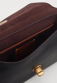 Intérieur d'un sac en cuir noir avec une doublure en suede bordeaux, quincaillerie dorée et une petite poche pour carte marron. Design minimaliste avec une texture lisse.