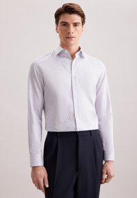 Jonge man in een wit en blauw verticaal gestreept overhemd met knopen en donker marineblauwe broek, staand tegen een effen lichte achtergrond.