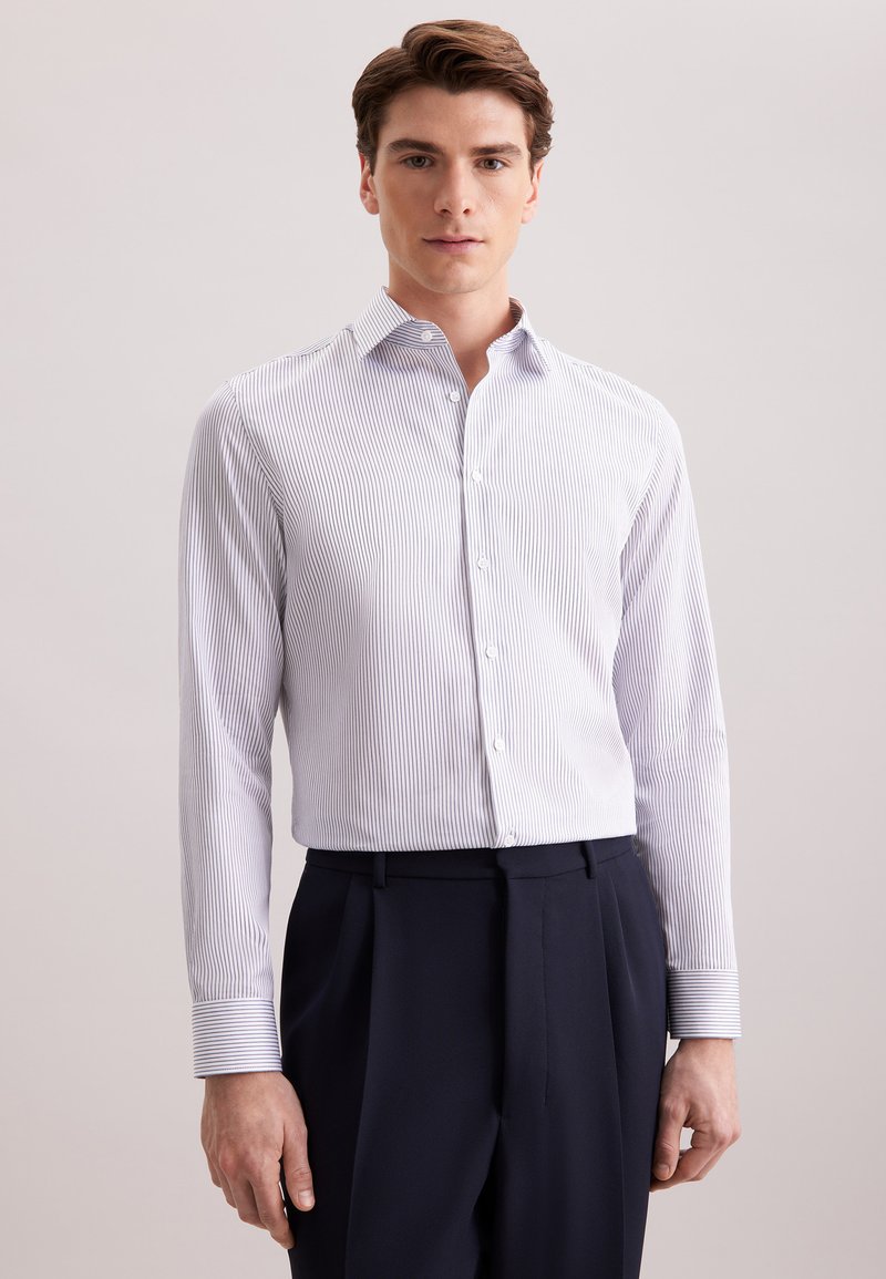 Jonge man in een wit en blauw verticaal gestreept overhemd met knopen en donker marineblauwe broek, staand tegen een effen lichte achtergrond.