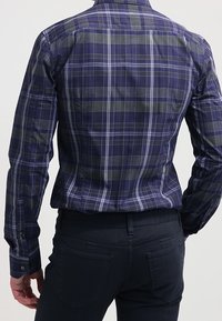 Hombre con una camisa de cuadros azul y verde de manga larga metida dentro de unos pantalones oscuros, de pie con la espalda hacia adelante contra un fondo sencillo.