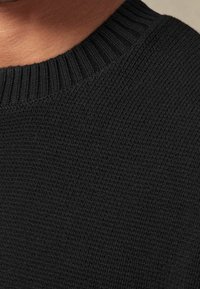 Schwarzer gestrickter Pullover mit einem gerippten Halsausschnitt und einer strukturierten Oberfläche, die das nahtlose, weiche Material und das schlichte Design hervorhebt.