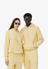 Sudaderas amarillas con capucha y bolsillos tipo canguro, pantalones a juego y logotipos discretos. Ambos modelos llevan gafas de sol; textura suave, corte relajado.