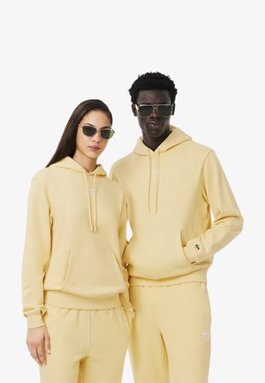 Sudaderas amarillas con capucha y bolsillos tipo canguro, pantalones a juego y logotipos discretos. Ambos modelos llevan gafas de sol; textura suave, corte relajado.