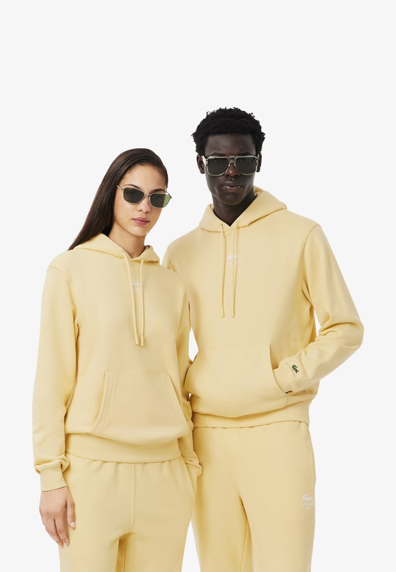 Sudaderas amarillas con capucha y bolsillos tipo canguro, pantalones a juego y logotipos discretos. Ambos modelos llevan gafas de sol; textura suave, corte relajado.