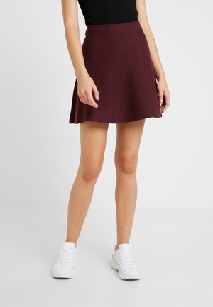 Femme portant une jupe trapèze bordeaux foncé, un haut sans manches noir et des baskets blanches, debout devant un fond clair uni.