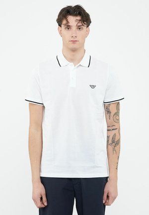 Joven con cabello corto y oscuro, de pie de frente, vistiendo una camisa polo blanca con ribetes negros y pantalones azul marino, con tatuajes visibles en el brazo derecho.