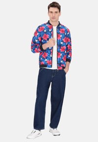 Blauwe bomberjack met een levendig roze en wit bloemenpatroon, zwarte manchetten en kraag. Gecombineerd met donkere spijkerbroeken en witte sneakers.
