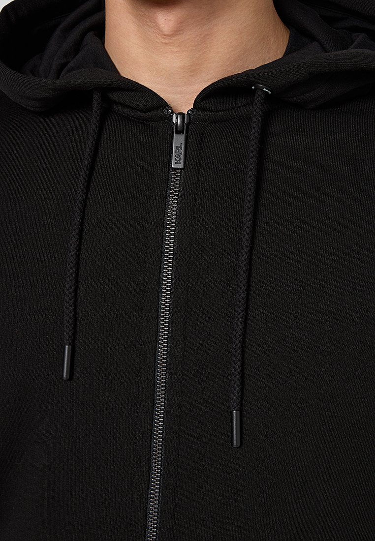 Sudadera negra con cremallera, con una superficie texturizada, cordones ajustables y una cremallera metálica que presenta un tirador de marca.