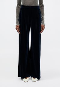 Pantalons en velours bleu marine à jambes larges, avec une finition texturée, dotés d'un devant plissé et d'une taille ajustée, associés à des chaussures blanches à bout pointu.