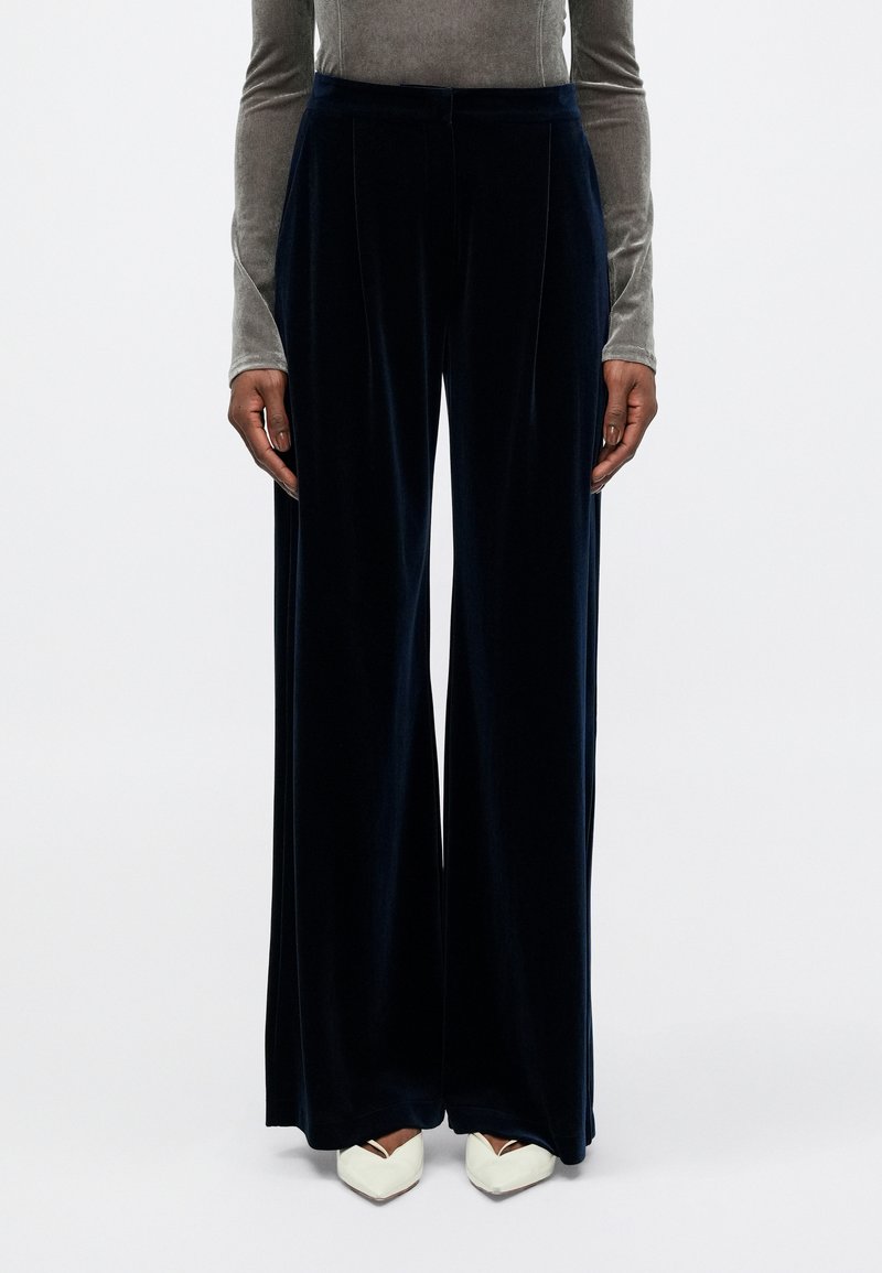 Pantalons en velours bleu marine à jambes larges, avec une finition texturée, dotés d'un devant plissé et d'une taille ajustée, associés à des chaussures blanches à bout pointu.