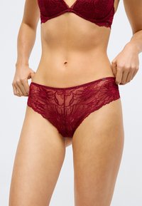 Vrouw in een rode kanten bh en bijpassende kanten slip, die haar slip aanpast bij de heupen tegen een effen achtergrond.