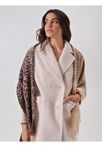 Femme aux cheveux bruns portant un manteau double boutonnage crème et un châle à motif marron et beige, regardant sur le côté.