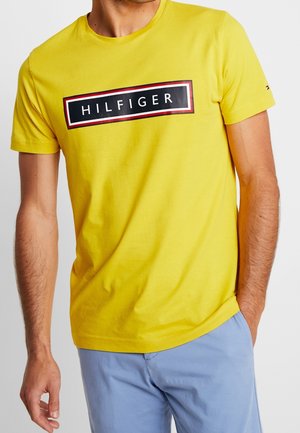 Maglietta di cotone giallo con un logo rettangolare nero che riporta la parola "HILFIGER" e dettagli di bordo rossi. Maniche corte e scollo a girocollo.