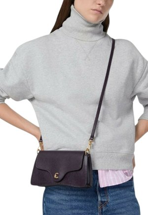 Borsa a tracolla in pelle color viola scuro con dettagli dorati, dotata di chiusura con patta e design curvo. Indossata sopra una felpa grigia.