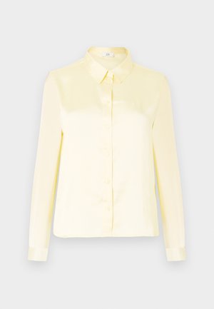Hosszú ujjú, világos sárga selyem blúz gombos elejével és klasszikus gallérral sima háttér előtt.