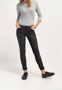 Vaqueros skinny negros con detalles horizontales con cremalleras en las rodillas, combinados con una camiseta polo de manga larga gris y zapatillas negras.