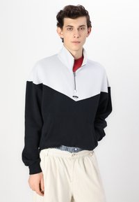 Svart och ljusgrå tröja med dragkedja vid halsen, med en kontrasterande chevron-design och en ficka framtill. Mjuk bomullsblandning.