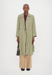 Trench coat verde chiaro con una vestibilità rilassata, con dettagli a bottoni, indossato sopra un polo bordeaux. Pantaloni beige larghi e scarpe nere slip-on completano l'outfit.