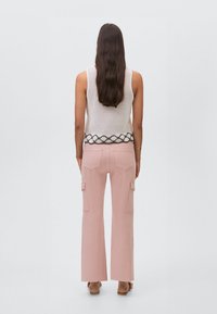 Žena s dlouhými tmavými vlasy, oblečená v bezrukávovém bílém pleteném topu s vzorovaným okrajem a růžových širokých cargo kalhotách, stojící na bílém pozadí.