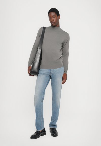 Gray turtleneck sweater, light blue denim jeans, black leather shoes, and a black shoulder bag. Model stands against a white backdrop.