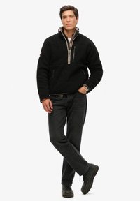 Czarny polarowy pulower z pół-zamkiem, z kieszenią na zamek błyskawiczny na klatce piersiowej, beżowymi akcentami i luźnym fasonem. Noszony z czarnymi dżinsami i butami.