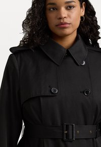 Svart trench coat med dubbelknäppt design, stor krage, två knappar, bälte i midjan och slät tygstruktur.