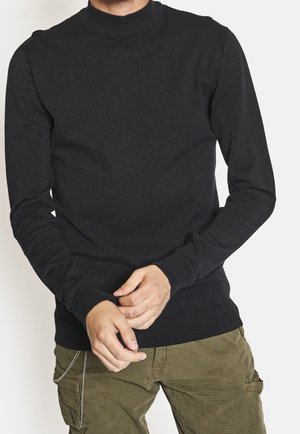 Man wearing a black long-sleeve turtleneck sweater and olive green cargo pants with a chain accessory.