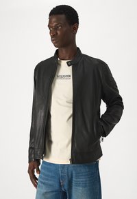 Giacca in pelle nera con colletto rialzato, chiusura lampo frontale, dettagli a coste sulle spalle e tasche laterali, abbinata a una maglietta beige chiaro.