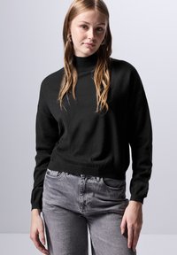 Maglione a collo alto nero, con texture e maniche lunghe, abbinato a jeans alti grigi chiari. Design semplice e casual senza motivi.