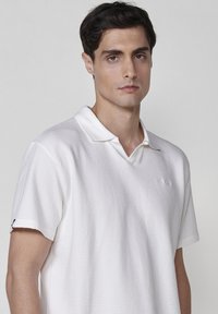 Vit kortärmad polo med texturerad tyg, med en klassisk krage och en liten logotyp på bröstet.