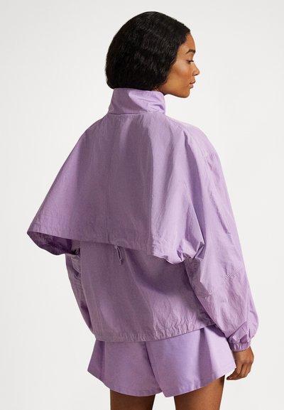 Γυναίκα που φοράει ένα φαρδύ, ανοιχτό μοβ μπουφάν με ψηλό γιακά και ταιριαστά σορτς, στέκεται πλευρικά μπροστά σε μια απλή φόντο.