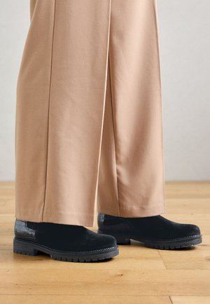 Bottines noires avec des sections en daim et en cuir verni, dotées d'une semelle épaisse et d'un dessus texturé, associées à un pantalon large beige.