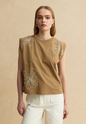 Frau trägt ein ärmelloses, beigefarbenes Oberteil mit weißer Blumenstickerei und weiße Hose, steht vor einem beigen Hintergrund.