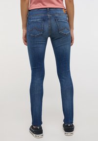 Jeans skinny de mezclilla azul con un lavado medio, que presentan bolsillos traseros con costuras curvas y una etiqueta de cuero en la cinturilla.