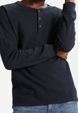 Osoba ubrana w czarną koszulę henley z długim rękawem i trzema guzikami, z założonymi ramionami, w połączeniu z niebieskimi dżinsami na jednolitym tle.