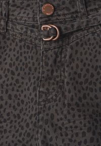 Close-up of dark gray fabric with black irregular spots, metal button with text, belt loop threaded through a bronze D-ring on patterned pants.