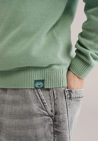 Pull en maille vert clair avec poignets et ourlet côtelés, portant une petite étiquette verte. Associé à un pantalon en denim gris.