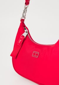 Sac à épaule en nylon rouge avec une texture lisse, fermeture éclair argentée et détail de logo. Dispose d'une bandoulière amovible et d'une forme arrondie.