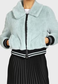 Giacca bomber in pelliccia sintetica azzurro chiaro con chiusura frontale a zip, polsini e orlo a costine neri con dettagli a strisce bianche, abbinata a una gonna nera plissettata.